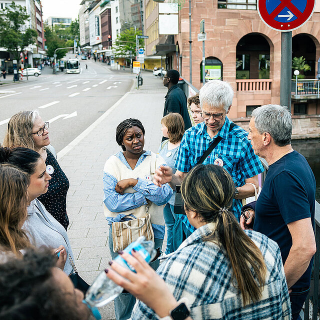 Ein Mann mit blau kariertem Hemd hält ein Tablet in der Hand und spricht. Um ihn stehen neun Personen und hören aufmerksam zu. - copyright:Stadt Pforzheim