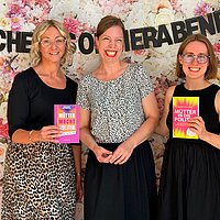 Drei Personen mit Büchern in der Hand vor einer Blumenwand.