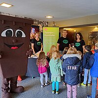 Eine Bio-Tonne als Maskottchen steht an einem Stand der Abfallwirtschaft. Davor stehen Kinder.