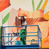 Künstlerin auf einem Scherenlift vor einer bunt bemalten Hausfassade
