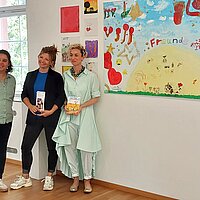 Drei Frauen stehen neben einem Bild, das von Kindern gemalt wurde. Auf dem Bild ist der Titel "Freunde" zu lesen.