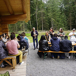 An zwei Tischen sitzen ungefähr 20 Personen, ein paar Personen stehen aus. Sie sind am Essen. Die Tische stehen vor einer Blockhütte am Waldrand. - copyright:Stadt Pforzheim