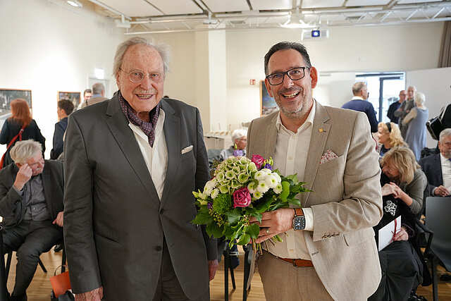 Zwei Männer im Anzug stehen vor vielen Menschen und schauen lächelnd in die Kamera. Ein Mann hält einen Blumenstrauß.