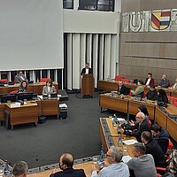 Mehrere Menschen sitzen in einem Sitzungssaal. Am Rednerpult steht ein Mann im Anzug.