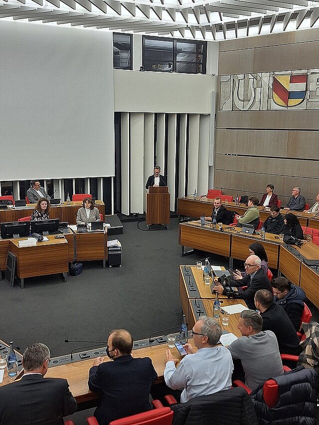 Mehrere Menschen sitzen in einem Sitzungssaal. Am Rednerpult steht ein Mann im Anzug.