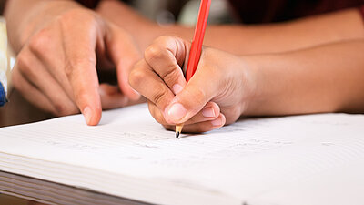 Großaufnahme: Kinderhand mit Stift und der Zeigefinger eines Erwachsenen. Symbolbild für Nachhilfe/Hausaufgabenbetreuung