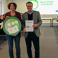 Auf dem Bild ist die stellvertretende Amtsleiterin der Stadtbibliothek, Marion Hekmann, zu sehen, die das Siegel aus der Hand von Staatssekretär Volker Schebesta, Ministerium für Kultus, Jugend und Sport, überreicht bekommt.