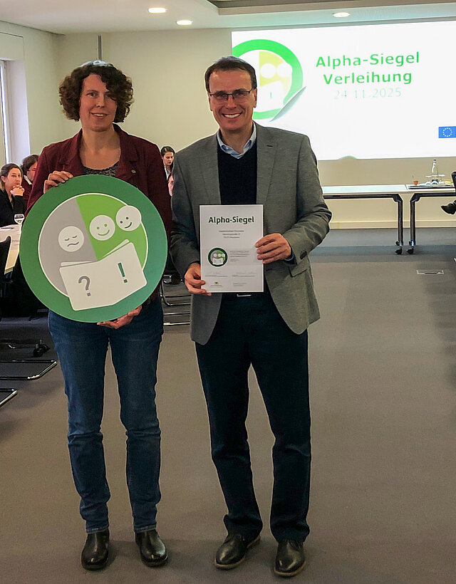 Auf dem Bild ist die stellvertretende Amtsleiterin der Stadtbibliothek, Marion Hekmann, zu sehen, die das Siegel aus der Hand von Staatssekretär Volker Schebesta, Ministerium für Kultus, Jugend und Sport, überreicht bekommt.