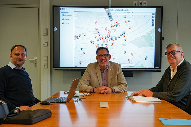 Baubürgermeister Tobias Volle (Mitte), Uwe Weiss, Leiter der Abteilung Straßenneubau und Verkehrstechnik im Grünflächen- und Tiefbauamt (rechts) und Johannes Halvaci, Leiter der Koordinierungsstelle, stellten die neue Baustellenübersicht der Stadt Pforzheim vor.  Sie sitzen vor einem großen Bildschirm auf dem die Baustellen auf einer Karte gezeigt werden.