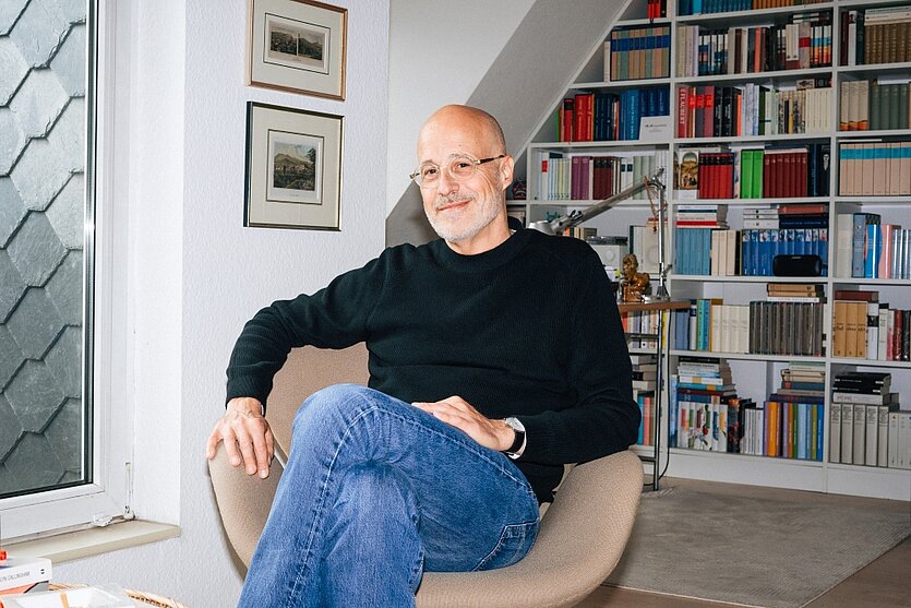 Ein Mann mit Brille und sehr kurzen grauen Haaren sitzt auf einem Stuhl vor einer Bücherwand. - copyright: Lea Sofia Fichtner - Ersteller: Lea Sofia Fichtner