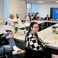 Mehrere Personen sitzen an Tischen in einer Fortbildungs-Situation, im Hintergrund ist ein eine Leinwand zu sehen.