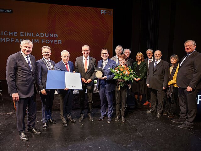 Eine Gruppe von Menschen stehen im Halbkreis und halten einen Blumenstrauß, eine Urkunde und eine Medaille.