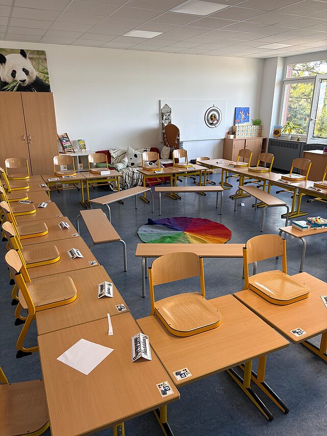 Das Bild zeigt einen Grundschulklassenraum, die Tische sind U-förmig angeordnet, die Stühle sind aufgestuhlt. In der Mitte des U stehen kreisförmig angeordnete Bänke.