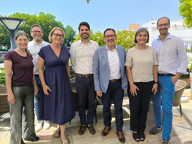 Sieben Personen auf einem Gruppenfoto, im Hintergrund ist ein blauer Himmel zu erkennen.