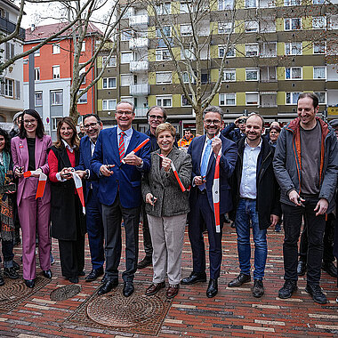 Viele Personen stehen nebeneinander auf einem Platz und halten je ein Stück rot-weißes Absperrband in den Händen