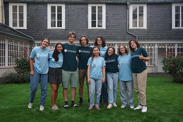 Farbfoto: 9 Schülerinnen und Schüler mit "Experiment"-T-Shirts auf einer Wiese vor einem Haus