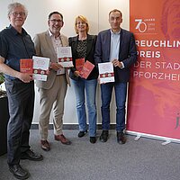 Vier Personen stehen vor einem orangenen Banner und halten Bücher in den Händen.