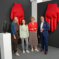 Kuratorin Tanja Solombrino, Mirijam Babel, wissenschaftliche Mitarbeiterin im Kulturamt sowie Claudia Baumbusch, Stellvertretende Leitung Kulturamt und Kulturbürgermeister Tobias Volle stehen vor einem Kunstwerk der Ausstellung Ortszeit und lächeln in die Kamera.