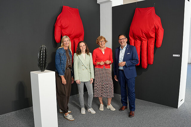 Kuratorin Tanja Solombrino, Mirijam Babel, wissenschaftliche Mitarbeiterin im Kulturamt sowie Claudia Baumbusch, Stellvertretende Leitung Kulturamt und Kulturbürgermeister Tobias Volle stehen vor einem Kunstwerk der Ausstellung Ortszeit und lächeln in die Kamera.