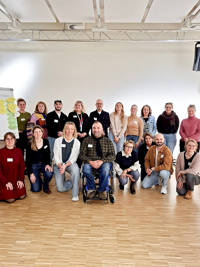 Eine Gruppe von Menschen steht, beziehungsweise sitzt in einem Raum. Neben ihnen ist ein Flipchart zu sehen.