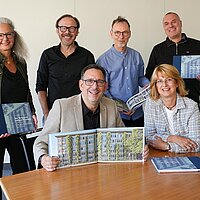 Sechs Personen stehen und sitzen an einem Tisch und präsentieren ein Buch