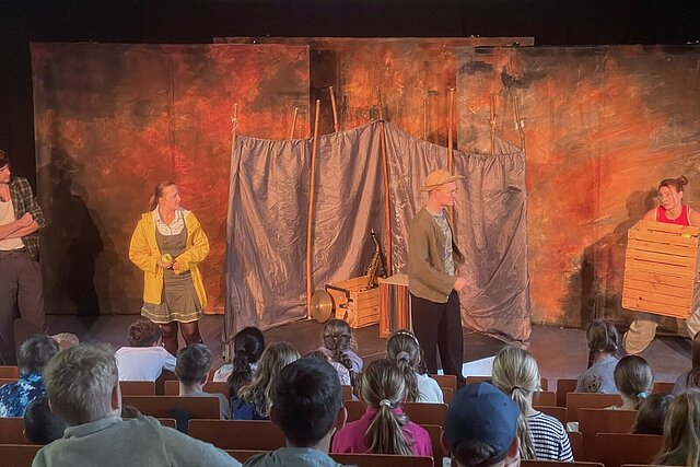 Im Hintergrund ist eine Theaterbühne zu sehen, im Vordergrund sitzen mehrere Schüler.