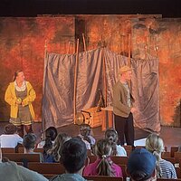 Im Hintergrund ist eine Theaterbühne zu sehen, im Vordergrund sitzen mehrere Schüler.