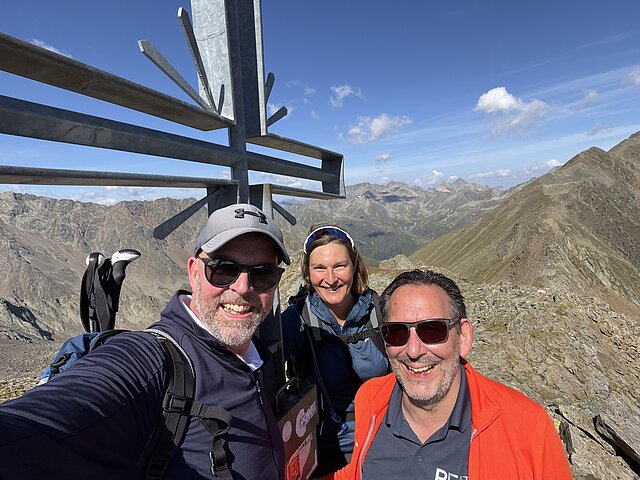 Bürgermeister Tobias Volle, Sonja Störzbach und Oliver Marc Heinz stehen vor einem Gipfelkreuz in den Tiroler Alpen.