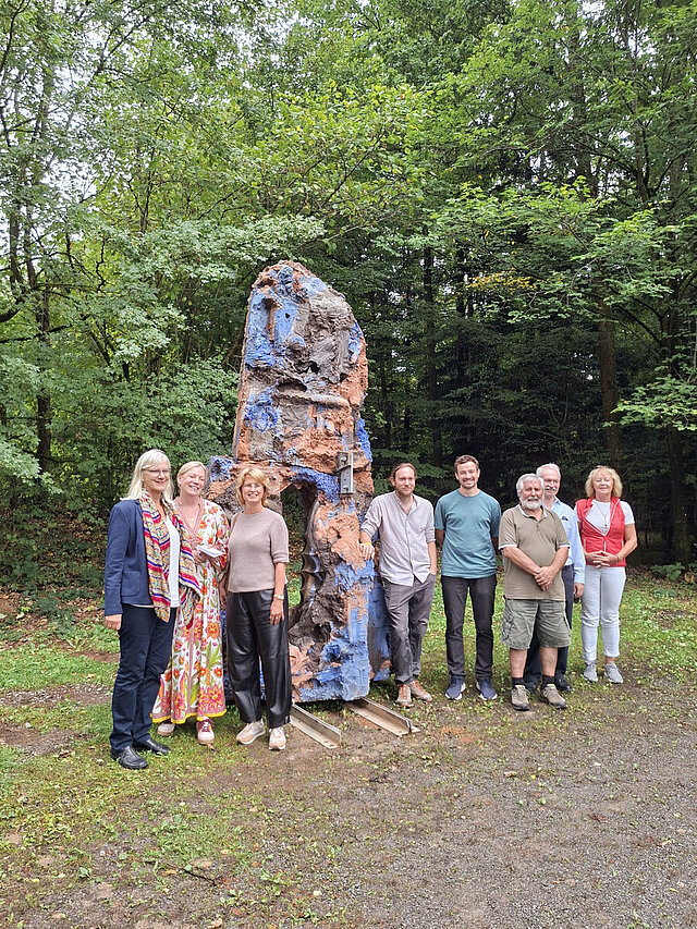 Acht Personen stehen vor einer Raben-Skulptur im Wald.