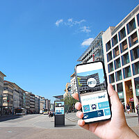 Das Bild zeigt eine digitale Stele und eine Hand mit einem Handy, auf dem die geplante Stadt-App zu sehen ist. 