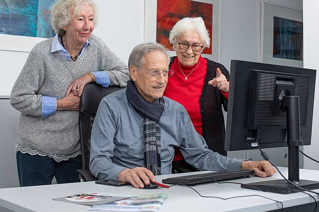 Drei Senioren sitzen und stehen am Schreibtisch und schauen in einen Computer