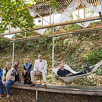 Fünf Personen sitzen auf einer Holzkonstruktion, eine Person liegt in einer Hängematte.