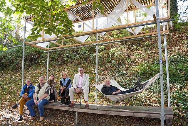 Fünf Personen sitzen auf einer Holzkonstruktion, eine Person liegt in einer Hängematte.