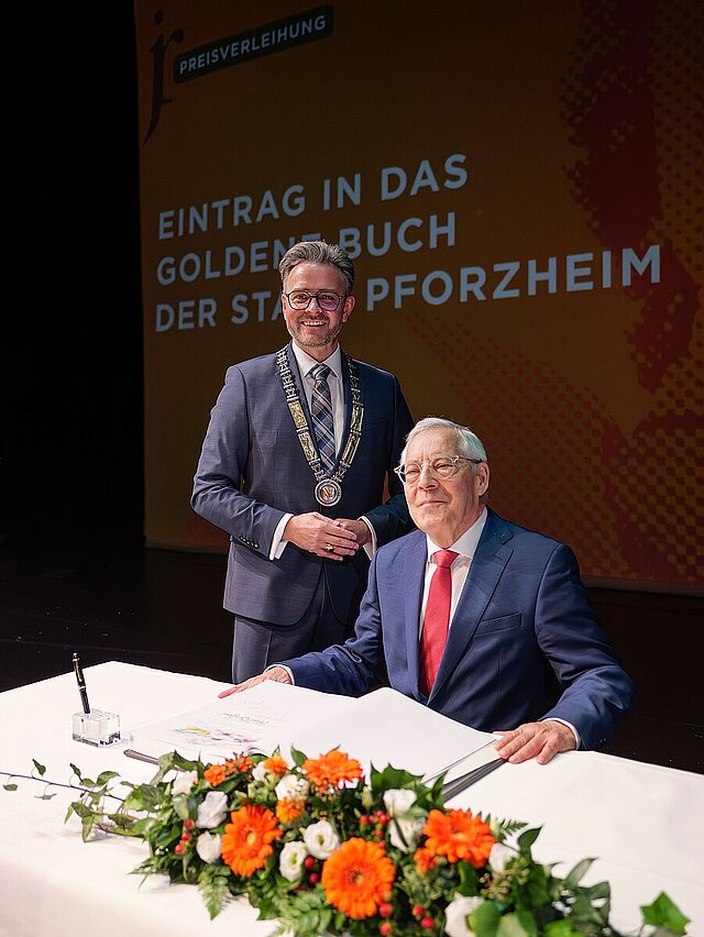 Ein Mann sitzt an einem Tisch, vor sich ein aufgeschlagenes Buch, ein Mann stehet hinter ihm. 