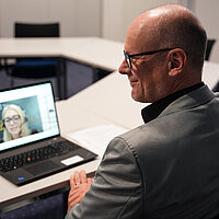 Das Bild zeigt Ersten Bürgermeister Dirk Büscher in einem Besprechungsraum, der an einem Laptop an einer Videokonferenz teilnimmt.  Zu sehen ist er von der Seite bzw. leicht von hinten. Er trägt ein graues Sakko und eine Brille und sitzt an einem Konferenztisch. Auf dem aufgeklappten Laptop ist das Bild einer zugeschalteten Gesprächspartnerin zu sehen.