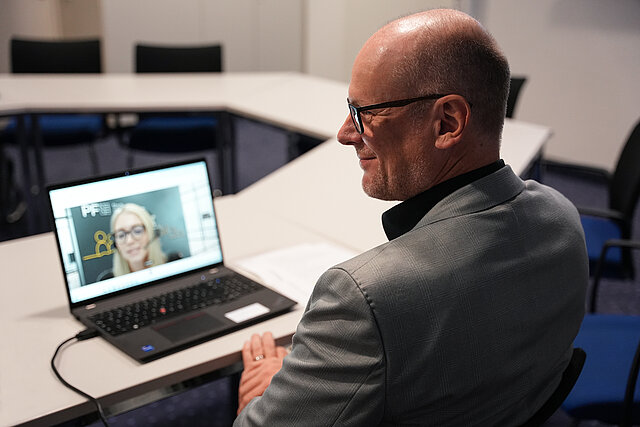 Das Bild zeigt Ersten Bürgermeister Dirk Büscher in einem Besprechungsraum, der an einem Laptop an einer Videokonferenz teilnimmt.  Zu sehen ist er von der Seite bzw. leicht von hinten. Er trägt ein graues Sakko und eine Brille und sitzt an einem Konferenztisch. Auf dem aufgeklappten Laptop ist das Bild einer zugeschalteten Gesprächspartnerin zu sehen.
