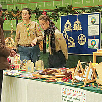 Zwei Frauen in Pfadfinderuniform hinter einem Stand auf einem Weihnachtsmarkt.