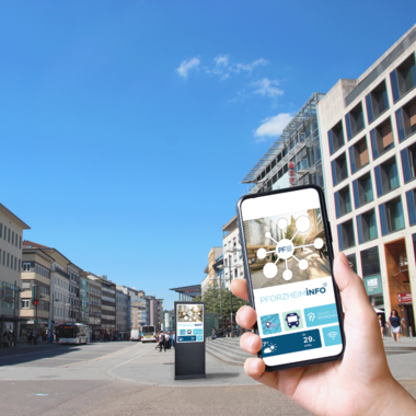 Die Pforzheimer Innenstadt. Im Vordergrund hebt eine Hand ein Handy, im Hintergrund ist eine Informationsstele zu sehen