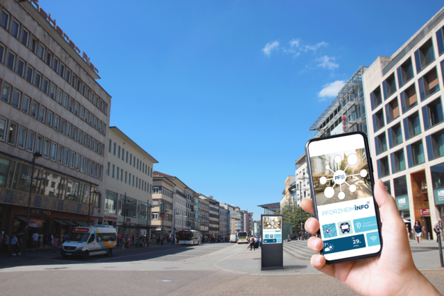 Die Pforzheimer Innenstadt. Im Vordergrund hebt eine Hand ein Handy, im Hintergrund ist eine Informationsstele zu sehen