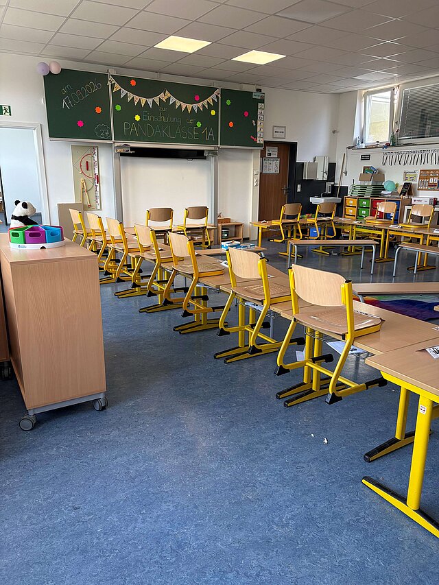 Das Bild zeigt einen Grundschulklassenraum mit Blick auf die Tafel. Die Tische stehen in einem Hufeisen, die Stühle sind aufgestuhlt.