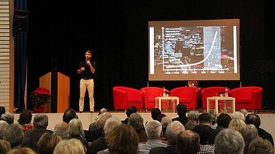Foto: Mann mit Mikro auf der Bühne, hält Präsentation