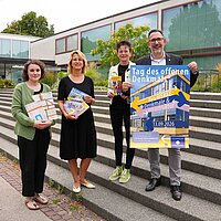 Vier Personen auf einer Treppe halten Plakate und stehen vor dem Schmuckmuseum.