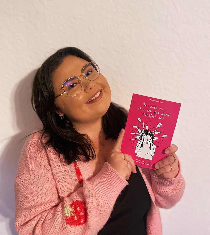 Frau mit Brille und rosa Cardigan hält ein Buch vor sich mit dem Text "ist halt so .. aber ist das denn wirklich so?" - copyright: Annie-Lauren Baur - Ersteller: Annie-Lauren Baur