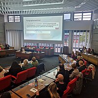 Viele Menschen sitzen in einem Sitzungssaal und schauen auf eine Präsentation