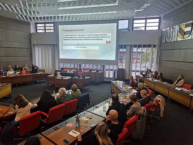 Viele Menschen sitzen in einem Sitzungssaal und schauen auf eine Präsentation