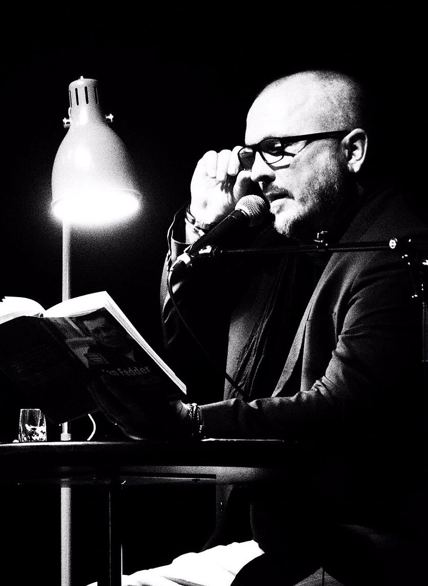 Schwarz/weiß Fotografie von Tim Pröse. Er sitzt an einem Tisch und liest ein Buch. - copyright: Paul Flemming für HEYNE - Ersteller: Paul Flemming für HEYNE