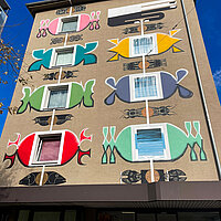Eine Wandgemälde an einer Hausfassade mit einem bunten Glücksarmband