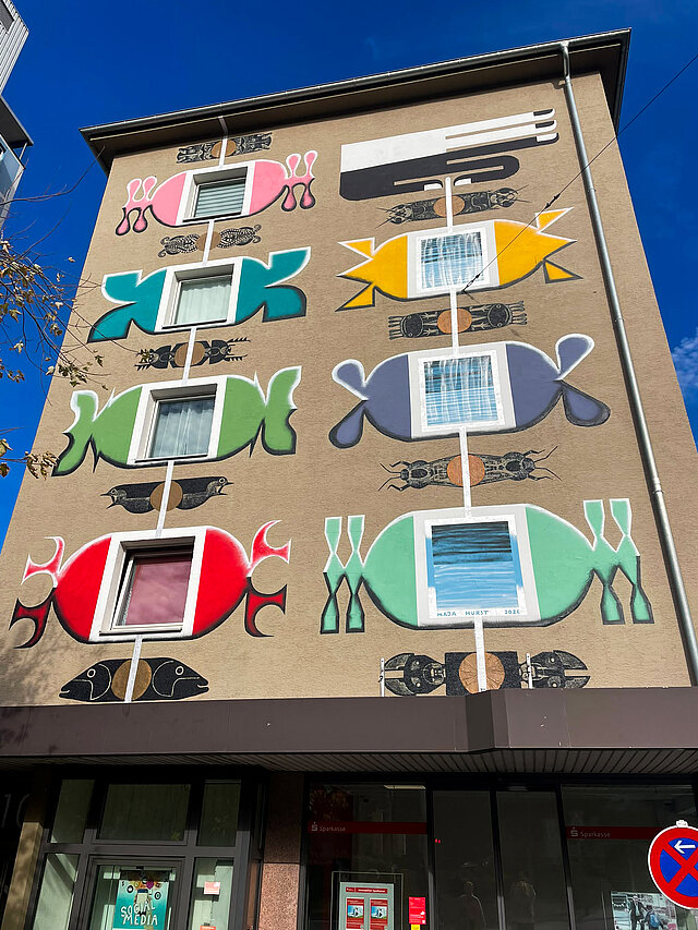 Eine Wandgemälde an einer Hausfassade mit einem bunten Glücksarmband