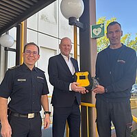 Feuerwehrkommandant Sebastian Fischer, Erster Bürgermeister Dirk Büscher und Notfallsanitäter sowie Praxisanleiter Stephan Bürkle (von links) stellen den neuen, öffentlich zugänglichen AED an der Hauptfeuerwache vor. Sie stehen dabei vor dem Eingang zur Hauptfeuerwache.