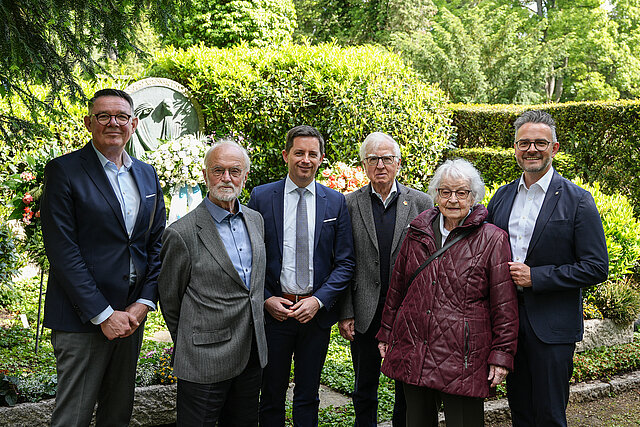 Gruppenfoto bei einer Kranzniederlegung auf einem Friedhof: Fünf Personen stehen gemeinsam vor einem begrünten Hintergrund mit Blumen und einem Gedenkstein. Oberbürgermeister Peter Boch steht rechts mit einem dunklen Anzug. Neben ihm befinden sich die Witwe von Werner Wild, Erika Wild, und weitere Männer in Anzügen. Die Gruppe blickt in die Kamera. Im Hintergrund sind Blumenarrangements und gepflegte Hecken zu sehen.